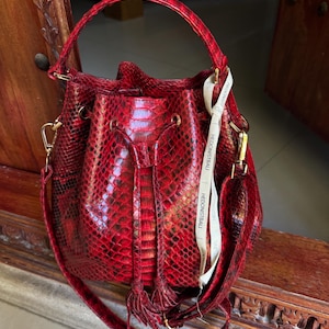May include: A red snakeskin bucket bag with a gold chain and a tassel. The bag has a long strap and a label that reads "Hedonistbali".
