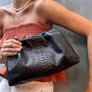 Könnte beinhalten: Eine schwarze Clutch aus Schlangenleder mit gerafftem, plissiertem Design. Die Tasche wird von einer Person gehalten, die ein rostfarbenes Oberteil und eine weiße Jeans trägt.