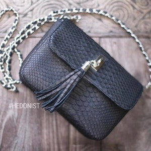 May include: A black snakeskin crossbody bag with a silver lock and a black tassel. The bag has a silver chain strap and the hashtag #HEDONIST is visible in the image.