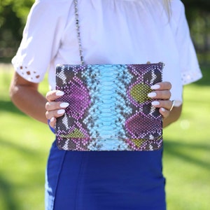 Op de afbeelding: Een rechthoekige clutch met een slangenprint in blauw, paars en geel. De tas heeft een zilveren kettingriem en een sluiting. De achtergrond is wazig, wat duidt op een buitenomgeving.