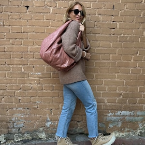 May include: A woman wearing a brown sweater, blue jeans, and beige hiking boots. She is carrying a pink leather handbag with a woven pattern.