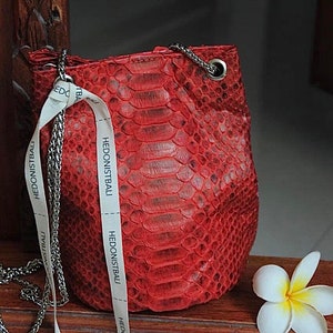 May include: A small red snakeskin handbag with a silver chain strap and white ribbon detailing. The ribbon has the text "HEDONISTBALI" printed on it.
