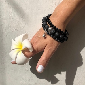 May include: A hand wearing three bracelets, two black lava stone bracelets and one black beaded bracelet with a silver charm. The hand is holding a white flower.