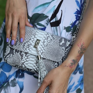 May include: A rectangular, gray and white snakeskin-patterned handbag with a silver chain strap and tassel. The bag has a silver clasp closure. The background includes a floral print dress with blue and white flowers.