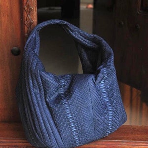 May include: A dark blue snakeskin handbag with a slouchy design. The bag has a single strap and is made of a textured material.