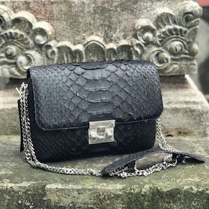 Op de afbeelding: Zwarte crossbody tas met een slangenleertextuur, zilveren ketting en zilveren sluiting. De rechthoekige tas heeft een klepsluiting. De tas ligt op een stenen oppervlak.