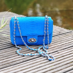 Könnte beinhalten: Eine kleine blaue Ledertasche mit einem silbernen Kettenriemen. Die Tasche hat einen Klappverschluss mit einem silbernen Verschluss.