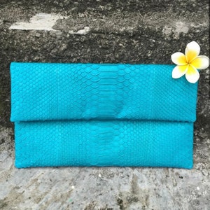 May include: A turquoise blue clutch purse made of snakeskin with a textured pattern. The clutch is closed and has a flap.