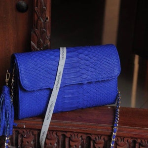 Könnte beinhalten: Eine kleine blaue Clutch aus Pythonleder mit einer goldenen Kettenkette und einem Quastenanhänger. Die Tasche hat ein weißes Etikett mit dem Text "Hedonistbali" darauf.