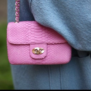 K&ouml;nnte beinhalten: Eine rosa Schultertasche mit Schlangenleder-Optik, einem silbernen Verschluss und einem silbernen Kettenriemen.