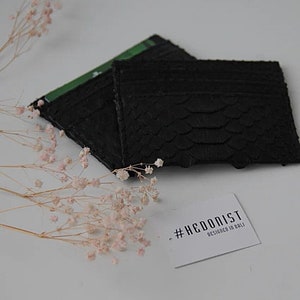 Puede incluir: Porta tarjetas de cuero negro con un patr&oacute;n de piel de serpiente. El porta tarjetas est&aacute; doblado y tiene una etiqueta blanca con el texto "#HEDONIST DESIGNED IN BALI".