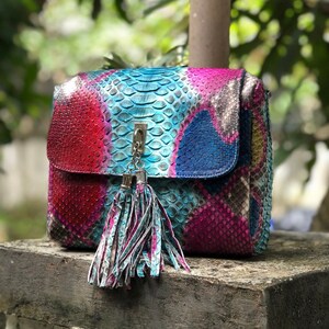 May include: A colorful, patterned handbag with a flap closure and a decorative tassel. The bag features a vibrant mix of red, blue, and purple hues, with a silver-tone clasp and tassel. The bag is made of a textured material, possibly leather.