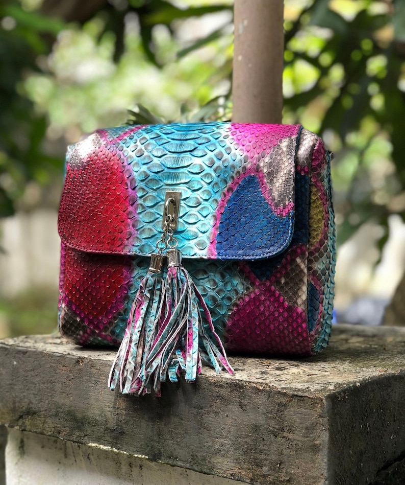 May include: A colorful, patterned handbag with a flap closure and a decorative tassel. The bag features a vibrant mix of red, blue, and purple hues, with a silver-tone clasp and tassel. The bag is made of a textured material, possibly leather.