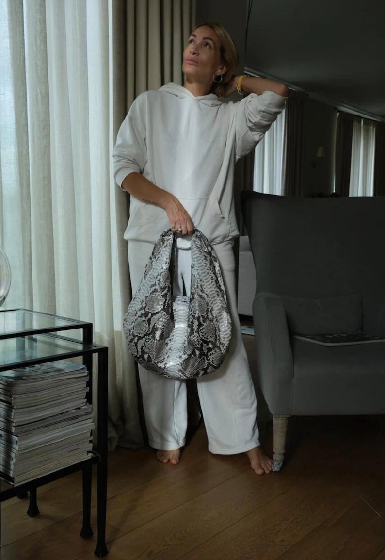 May include: A woman wearing a white hoodie and matching sweatpants is holding a large, gray and black snakeskin handbag. The bag has a unique, slouchy design and is made of a luxurious material.