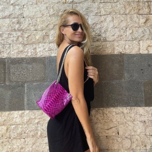 Op de afbeelding: Een vrouw in een zwarte, mouwloze jurk en zonnebril draagt een klein, felroze schlangenleren handtasje met een zilveren kettingband.