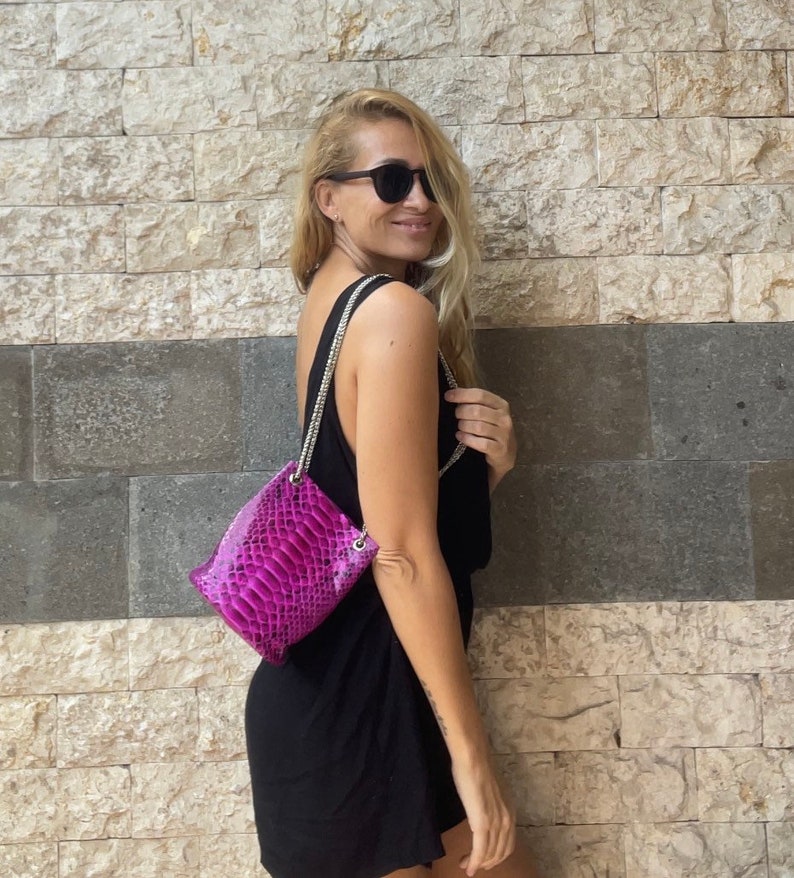 Op de afbeelding: Een vrouw in een zwarte, mouwloze jurk en zonnebril draagt een klein, felroze schlangenleren handtasje met een zilveren kettingband.