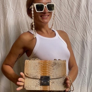 May include: A beige and brown snakeskin-patterned handbag with a gold chain strap and a gold lock closure.