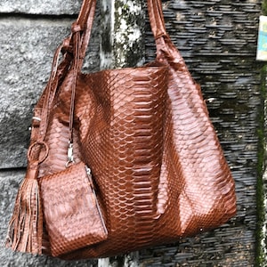 May include: A brown leather tote bag with a snakeskin pattern and a matching brown leather pouch with a tassel. The bag has a long strap and a zipper closure.