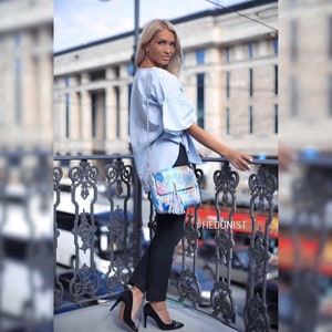 May include: A light blue blouse with ruffled sleeves and a patterned handbag with a tassel. The handbag has a silver chain strap. The image also shows black pants and black high heels. The hashtag #HEDONIST is visible.