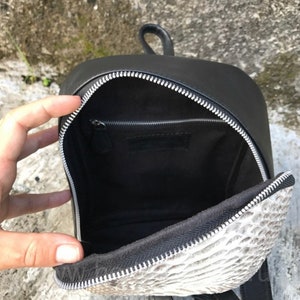 以下が含まれることがあります： ジッパー留めの小さな黒い革のバックパック。バックパックの内側は薄いグレーのヘビ柄で裏打ちされています。