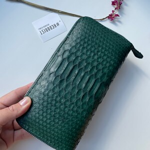 以下が含まれることがあります： ジッパー留めのダークグリーンのヘビ革財布。財布には、「#HECOONIST  DESIGN BY COONIST」というテキストが書かれた白いタグが付いています。