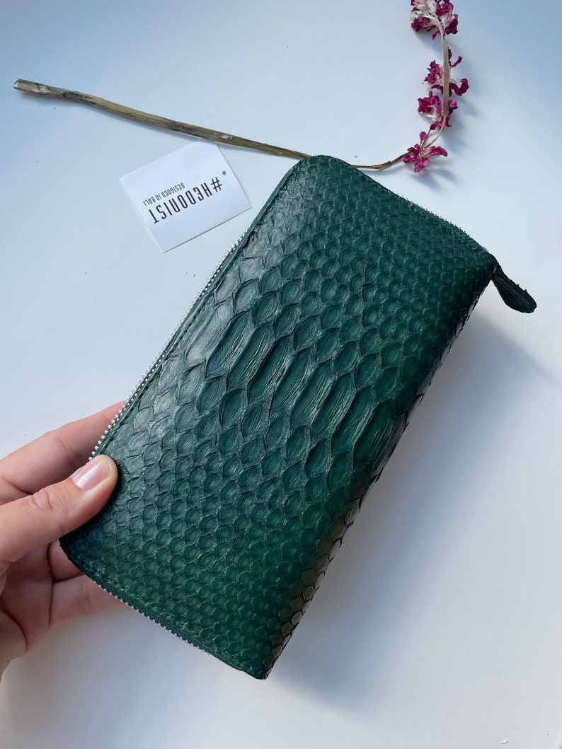 以下が含まれることがあります： ジッパー留めのダークグリーンのヘビ革財布。財布には、「#HECOONIST  DESIGN BY COONIST」というテキストが書かれた白いタグが付いています。