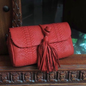 May include: A red leather clutch bag with a tassel. The bag has a snakeskin pattern and a knotted closure.