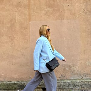 May include: A light blue blazer with a black crossbody bag and gray pants. The blazer has a classic design with a two-button closure and a notched lapel. The black bag has a chain strap. The pants are a relaxed fit. White sneakers complete the outfit.