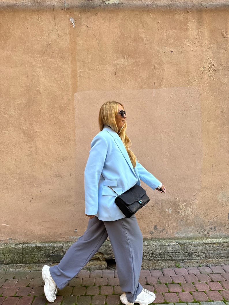 May include: A light blue blazer with a black crossbody bag and gray pants. The blazer has a classic design with a two-button closure and a notched lapel. The black bag has a chain strap. The pants are a relaxed fit. White sneakers complete the outfit.
