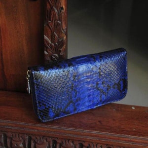 Op de afbeelding: Een blauwe slangenleren portemonnee met een ritssluiting. De portemonnee ligt op een houten oppervlak met ingewikkelde houtsnijwerk.