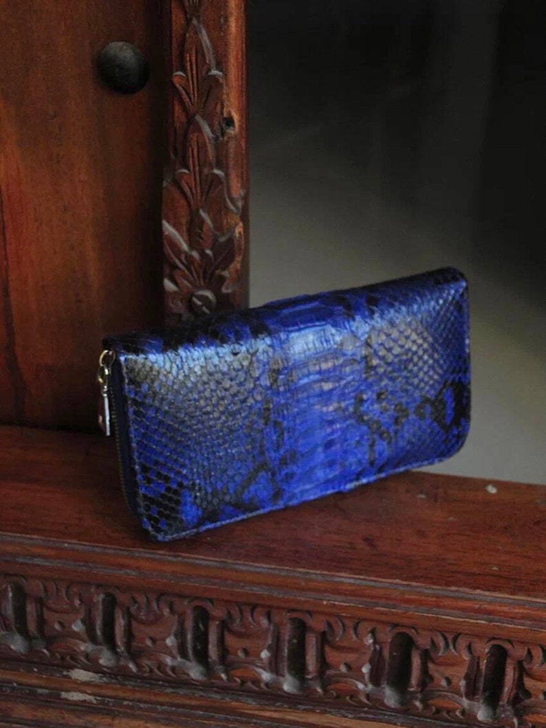 Op de afbeelding: Een blauwe slangenleren portemonnee met een ritssluiting. De portemonnee ligt op een houten oppervlak met ingewikkelde houtsnijwerk.