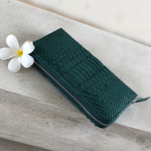 以下が含まれることがあります： ジッパー留めの濃い緑色の革財布。財布は、蛇革の質感のあるパターンです。