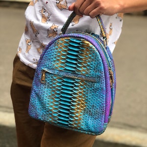 Könnte beinhalten: Ein blauer und goldener Schlangenleder-Rucksack mit Reißverschluss. Der Rucksack hat einen oberen Griff und verstellbare Träger.