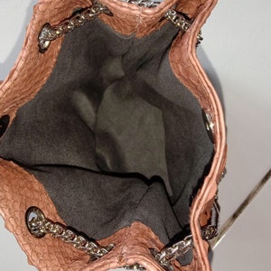May include: A light brown snakeskin bucket bag with a silver chain strap. The bag is open and the inside is lined with a dark grey fabric.