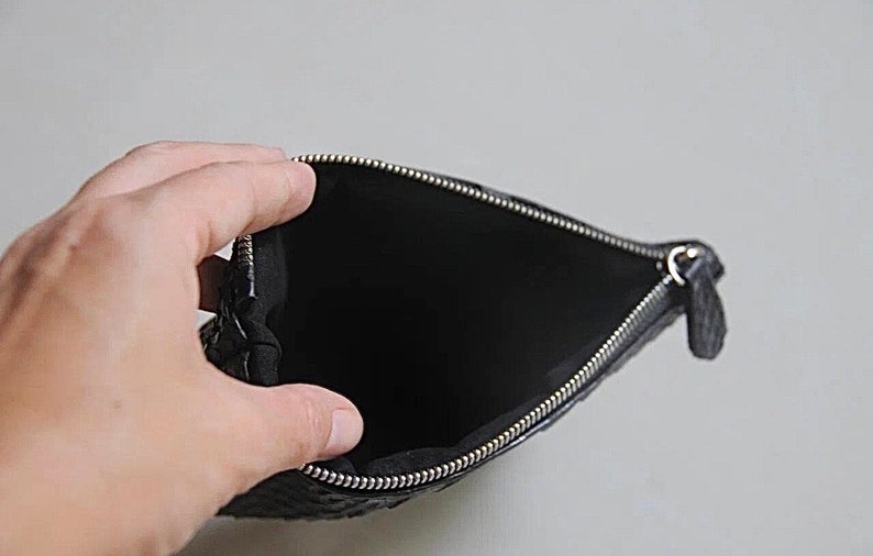 Może przedstawiać: Czarna sk&oacute;rzana kosmetyczka z srebrnym zamkiem błyskawicznym. Kosmetyczka jest otwarta i pusta.