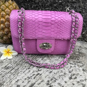 K&ouml;nnte beinhalten: Eine kleine, helllila Handtasche mit einer Kettenkette. Die Tasche ist aus Schlangenleder und hat einen silbernen Verschluss.