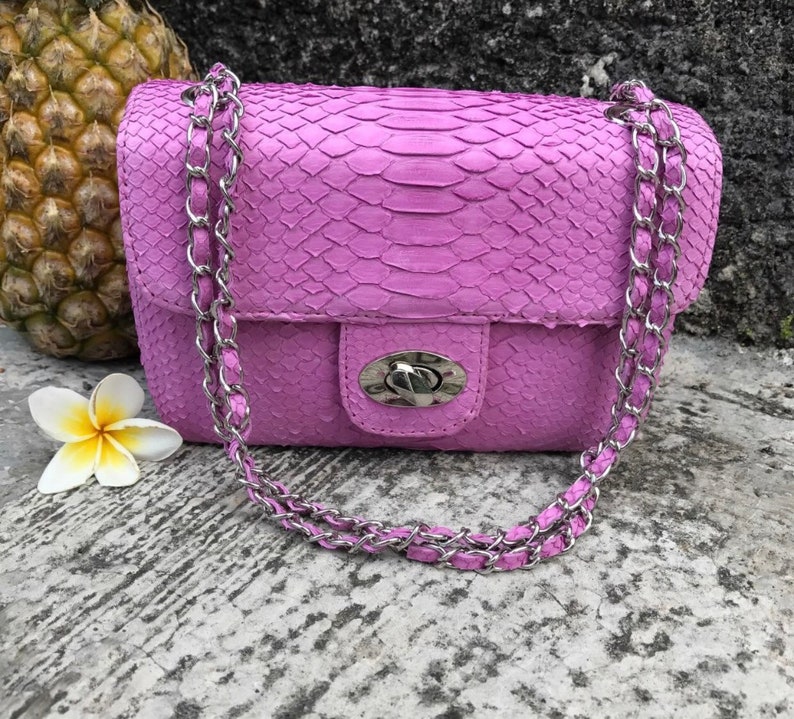 K&ouml;nnte beinhalten: Eine kleine, helllila Handtasche mit einer Kettenkette. Die Tasche ist aus Schlangenleder und hat einen silbernen Verschluss.