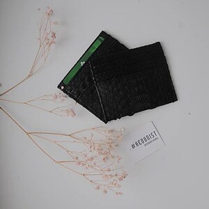 Puede incluir: Porta tarjetas de cuero negro con m&uacute;ltiples ranuras para tarjetas. El porta tarjetas est&aacute; hecho de cuero texturizado y tiene un dise&ntilde;o minimalista. El porta tarjetas est&aacute; sobre una superficie blanca con un peque&ntilde;o ramo de flores rosas.