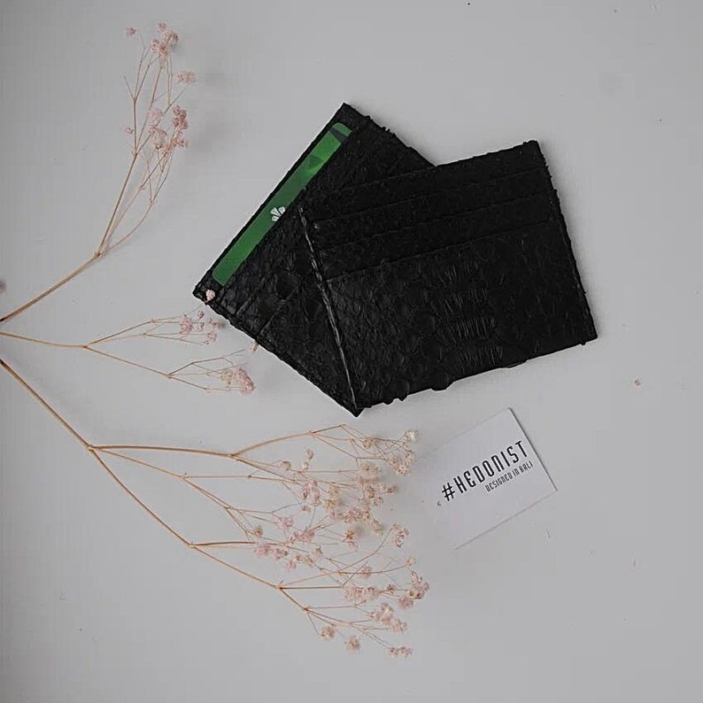 Puede incluir: Porta tarjetas de cuero negro con m&uacute;ltiples ranuras para tarjetas. El porta tarjetas est&aacute; hecho de cuero texturizado y tiene un dise&ntilde;o minimalista. El porta tarjetas est&aacute; sobre una superficie blanca con un peque&ntilde;o ramo de flores rosas.