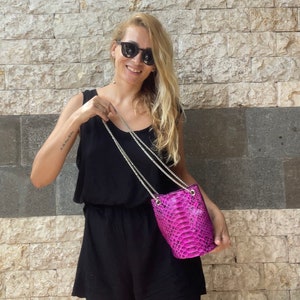 Op de afbeelding: Een vrouw in een zwarte, mouwloze jumpsuit en zonnebril houdt een kleine roze slangleren emmertassen met een zilveren kettingriem.