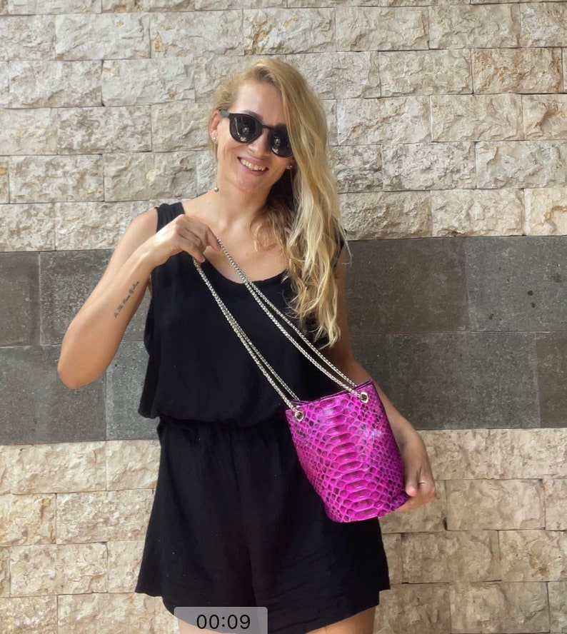 Op de afbeelding: Een vrouw in een zwarte, mouwloze jumpsuit en zonnebril houdt een kleine roze slangleren emmertassen met een zilveren kettingriem.