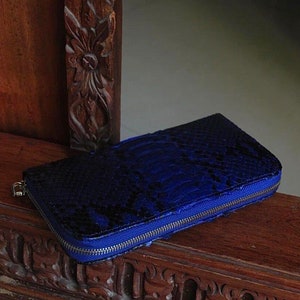 Op de afbeelding: Een blauwe slangenleren portemonnee met een ritssluiting. De portemonnee ligt op een houten oppervlak.