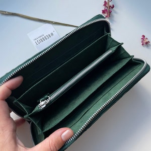 以下が含まれることがあります： ジッパー留めの濃い緑色の革財布。財布が開いており、複数のカードスロットとジッパー付きのコンパートメントが見えます。財布には「#REC00151  DESIGN BY KOOIST」というラベルが付いています。