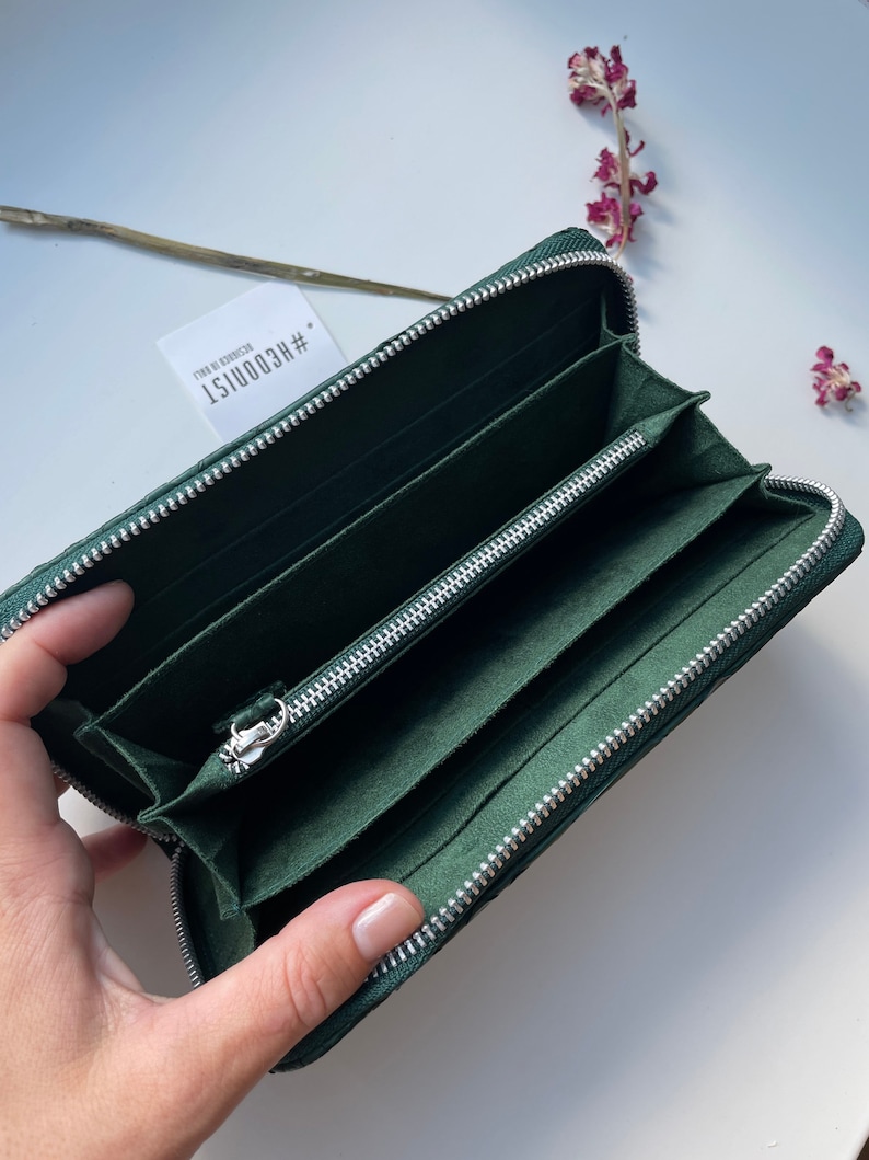 以下が含まれることがあります： ジッパー留めの濃い緑色の革財布。財布が開いており、複数のカードスロットとジッパー付きのコンパートメントが見えます。財布には「#REC00151  DESIGN BY KOOIST」というラベルが付いています。