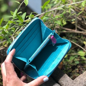 以下が含まれることがあります： ジッパー留めのターコイズブルーの財布。財布が開いており、内側は柔らかなブルーの生地で裏打ちされています。