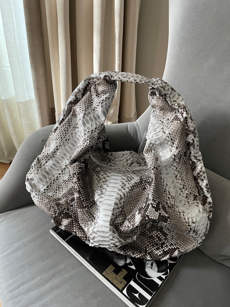 May include: A large, slouchy handbag made of snakeskin with a light brown and white pattern. The bag is sitting on a gray armchair with a magazine on top of it.