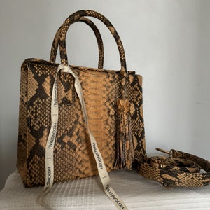 以下が含まれることがあります： トップハンドルと取り外し可能なショルダーストラップが付いた、小さく、茶色と黒のヘビ革製のハンドバッグ。バッグの正面にはタッセルと「Hedonistbali」と書かれたラベルが付いています。