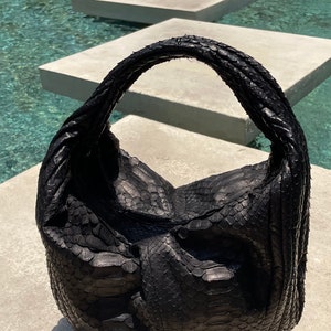 Może przedstawiać: Czarna skórzana torebka z wzorem skóry węża. Torebka ma jeden uchwyt i luźny, nieustrukturyzowany kształt.