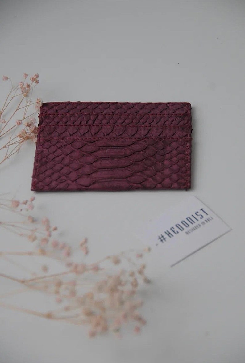 K&ouml;nnte beinhalten: Ein Kartenetui aus burgunderfarbenem Leder mit einem Schlangenhautpr&auml;gemuster. Das Kartenetui liegt auf einer wei&szlig;en Oberfl&auml;che mit einer Visitenkarte, auf der "#HEDONIST DESIGNED IN ITALY" steht.