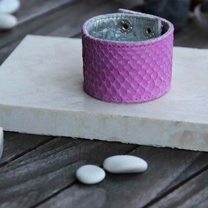 Pode incluir: Um bracelete de couro roxo com um padrão de pele de cobra. O bracelete está sobre uma superfície branca.